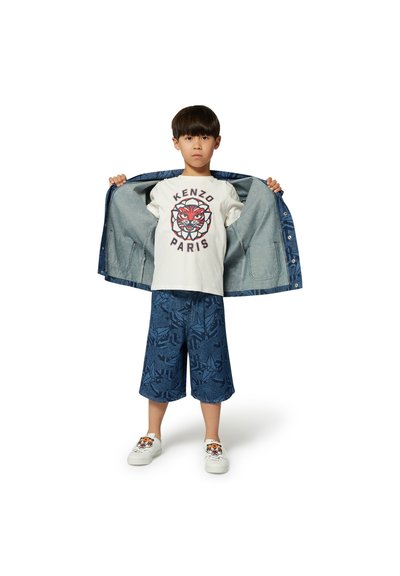 Spijkerjack, wit KENZO T-shirt met tijgerlogo, blauw patroonshorts en witte schoenen met tijgerdesign, alles gedragen door een kind tegen een neutrale achtergrond.