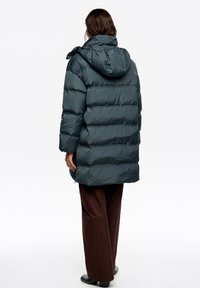 Zielona puchowa kurtka z poziomym pikowaniem i kapturem, zapinana na zamek błyskawiczny, z bocznymi kieszeniami, noszona na brązowych szerokich spodniach.