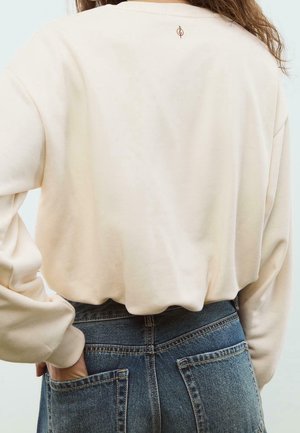 Persoon in beige shirt met lange mouwen met klein logo op de bovenrug, blauwe spijkerbroek, hand in achterzak gestoken.