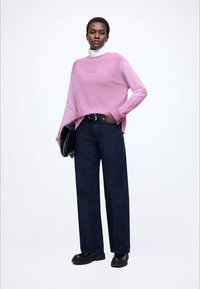 Ροζ πουλόβερ με στρογγυλή λαιμόκοψη και μακριά μανίκια, συνδυασμένο με σκούρο μπλε τζιν παντελόνι με φαρδιά πατζάκια. Το μοντέλο κρατάει μια μαύρη τσάντα χειρός.