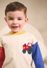 Maglietta di cotone con design multicolore: corpo crema, maniche rosse, accenti blu e un trattore rosso ricamato con ruote blu.