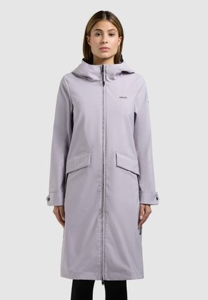 Femeie purtând o haină lungă gri deschis cu glugă, cu fermoar frontal și buzunare cu clapetă, stând pe fundal deschis simplu.