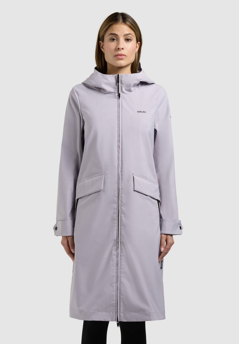 Femme portant un long manteau à capuche gris clair avec une fermeture éclair à l'avant et des poches à rabat, se tenant devant un fond uni clair.
