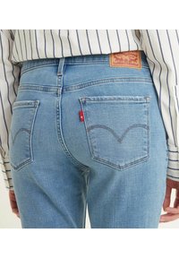 Vaqueros de mezclilla azul claro con cintura alta, bolsillos traseros curvados y una etiqueta de cuero con un logo en la cinturilla. Camiseta a rayas parcialmente visible.