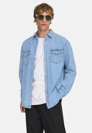 Ung man i ljusblå jeansskjorta över vit t-shirt, svarta byxor och gröntonade solglasögon, står med ena handen i fickan.