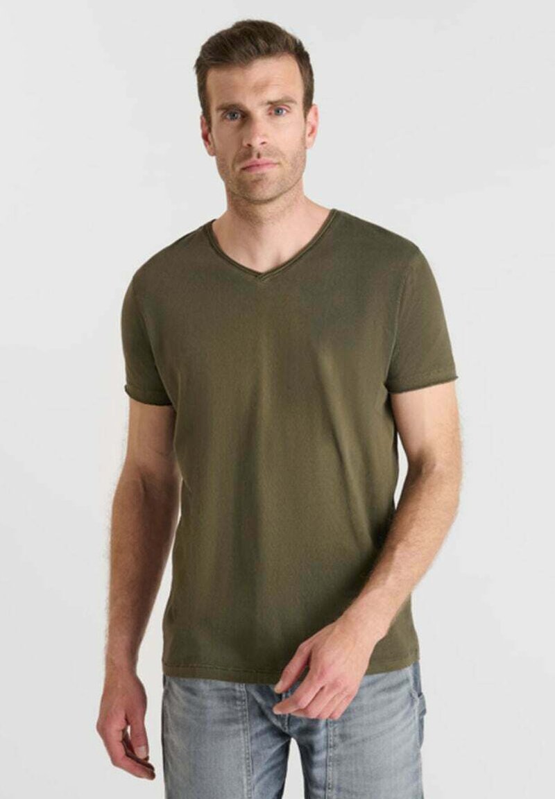 Homme portant un T-shirt à col en V à manches courtes de couleur vert olive et un jean bleu clair, se tenant devant un fond gris clair uni.