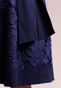 Laivastonsininen mekko, jossa on monimutkainen kukkakirjonta ja sileä satiinivartalo. Alahelma on vahvistettu tummansinisellä cuffilla, mikä tuo rakenteellista yksityiskohtaa.