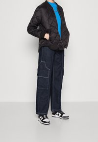 Schwarze gesteppte bomberjacke mit einem blauen Rundhalshemd, dunkelblauen Cargo-Jeans mit weißer Naht und schwarz-weißen Sneakers.