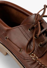 Zapato náutico de cuero marrón con ojales metálicos, costuras contrastantes y una plantilla texturizada. Los cordones están atados cuidadosamente en el lateral.