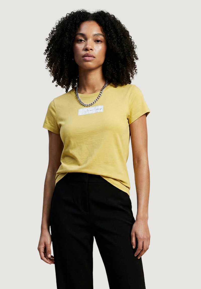Mujer con cabello rizado, vestida con una camiseta amarilla de manga corta y pantalones negros, de pie contra un fondo claro y liso.
