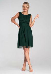 Grünes ärmelloses Kleid mit tailliertem Oberteil und schwingendem Rock. Verfügt über ein plissiertes Design und eine transparente Overlay. Getragen mit nudefarbenen Riemchen-Heels.