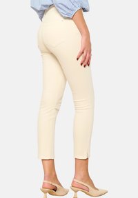 Jeans skiny beige con una textura suave, cintura media, bolsillos traseros y aberturas en los tobillos. Combinados con sandalias de tacón nude.
