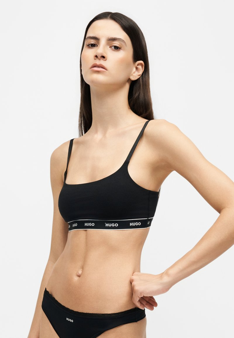 Femme aux longs cheveux foncés portant un soutien-gorge de sport noir marqué Hugo et un sous-vêtement noir assorti, debout avec une main sur la hanche.