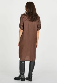 Rochie cămașă brown, până la genunchi, cu mâneci răsucite, dintr-un material transparent. Asortată cu cizme din piele neagră, până la genunchi, cu catarame.