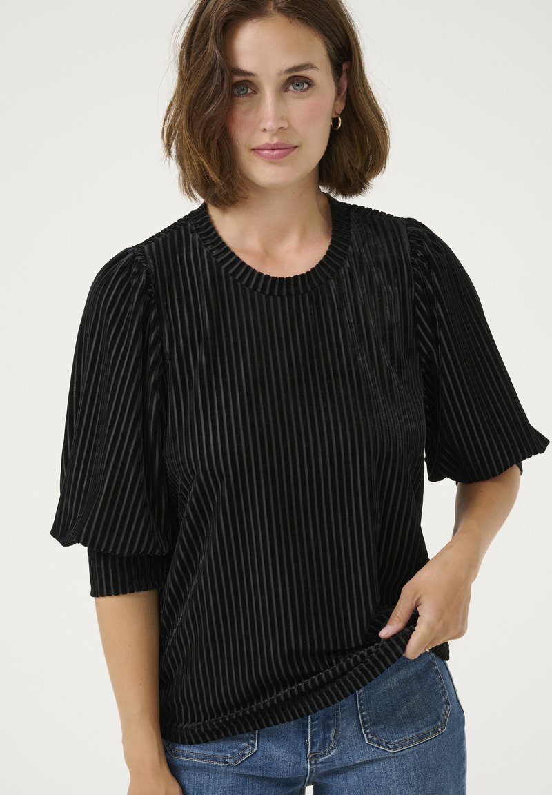 Top in velluto nero con pieghe verticali, scollatura rotonda e maniche corte a sbuffo. Abbinato a jeans in denim blu.