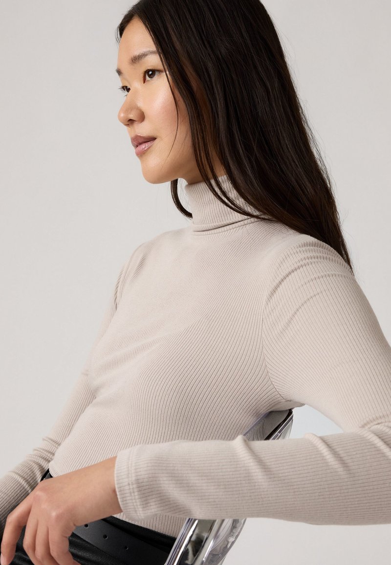 Beige geripptes Rollkragenoberteil mit langen Ärmeln. Besitzt ein figurnahes Design und eine weiche Textur, getragen mit einem schwarzen Gürtel, vor einem neutralen Hintergrund.