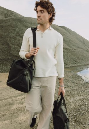 Ung mann med krøllete hår som går på grus, iført hvit genser og beige bukser, bærende to svarte vesker utendørs.