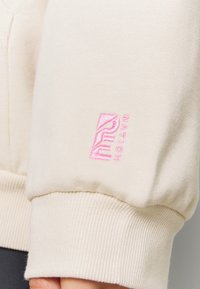 Manche de sweat-shirt blanc cassé avec poignet côtelé et logo brodé rose "PUMA NATION" près du poignet.