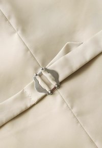 Ceinture en tissu beige avec une boucle métallique argentée à double ardillon sur un fond textile beige assorti.