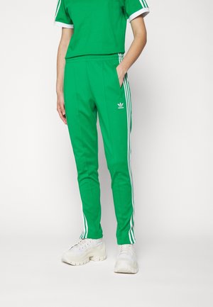 Personne portant un pantalon de survêtement Adidas vert avec des bandes blanches sur les côtés, associé à des baskets blanches épaisses et un haut vert assorti.