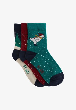 Trois paires de chaussettes : teal avec un bonhomme de neige patineur, bourgogne avec des accents blancs, et crème avec un orteil vert. Chacune présente des motifs ludiques.