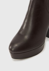 Σκούρο καφέ πλατφόρμα δερμάτινη μποτάκια αστραγάλου με στρογγυλεμένη μύτη και χοντρό ψηλό τακούνι σε λευκό φόντο.