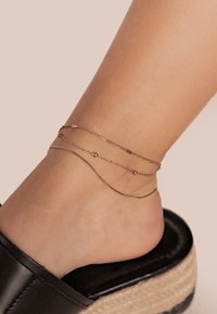 Pulsera de tobillo que presenta tres delicadas cadenas en tono dorado, incluyendo pequeñas cuentas y diseños de eslabones variados, llevada en un pie desnudo.