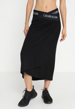 Frau trägt einen schwarzen asymmetrischen Midirock mit einem Taillenbund mit Calvin Klein Logo und schwarzen Sportschuhen, steht vor einem schlichten weißen Hintergrund.