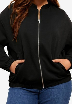 Femme portant une veste noire zippée avec les mains dans les poches et un jean bleu, montrée du cou aux hanches.