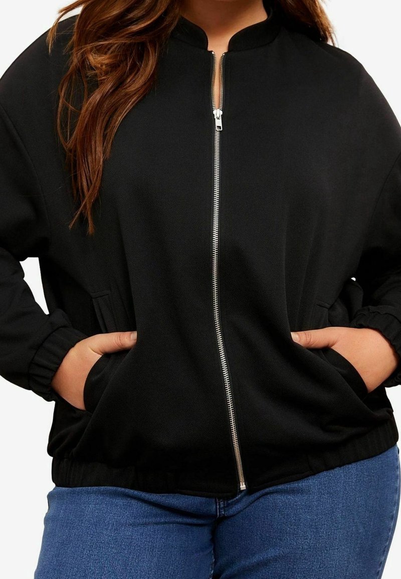 Femme portant une veste noire zippée avec les mains dans les poches et un jean bleu, montrée du cou aux hanches.