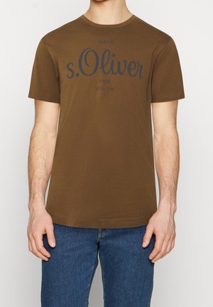 Mand iført brun t-shirt med korte ærmer med "s.Oliver" logo og blå jeans, stående mod en ensfarvet lys baggrund.