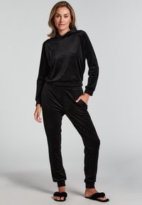 Musta veluurinen urheiluasu, jossa on huppari ja kapeat housut sivutaskuilla, yhdistettynä pörröisiin mustiin liukumasan kenkiin. Sujuva pinta.