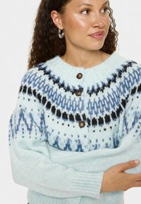Nainen, jolla on yllään vaaleansininen neulecardigan, jossa on sinimustalla kuviolla varustettu kaulus, napitettu etuosa ja hopeiset renkaakorvakorut, katsoo sivulle.
