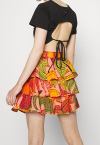 Černý zkrácený top s nařaseným zadním dílem a tkaničkou, kombinovaný s barevnou sukní s volány, která je zdobena živými botanickými potisky a s volánkovým lemem.