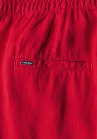 Röda tygshorts med elastisk midja. Har en liten, sydd ficka med en silvertagg märkt "Signet One." Mjuk textur.