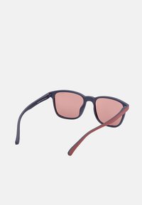 Gafas de sol con marco oscuro, forma rectangular y lentes con degradado en rosa. La patilla tiene un acento rojo en contraste. Acabado mate.