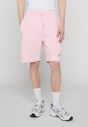 Persona che indossa pantaloncini rosa chiaro con coulisse e tasche laterali, calze bianche e scarpe da ginnastica New Balance bianche.