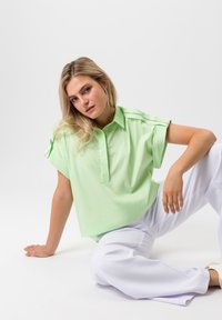 Lichtgroene oversized shirt met een kraag, korte opgerolde mouwen en een knoopsluiting, gecombineerd met witte wijde broek. Soepele textuur.