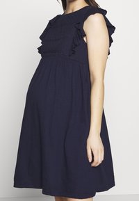 Granatowa sukienka ciążowa z falbaniastymi akcentami na ramionach i teksturowanym korpusem, luźny krój dla komfortu, długość do kolan, wykonana z miękkiego materiału.