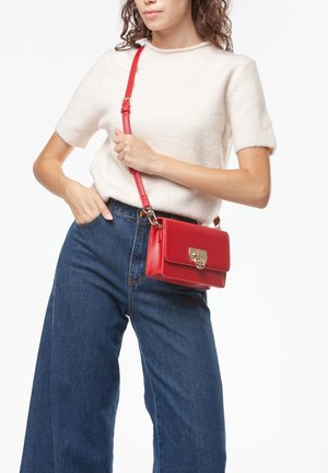 Vrouw met een wit, kort mouwsweater en blauwe spijkerbroek, die een rode leren schoudertas met gouden sluiting over haar torso draagt.