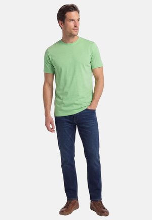 Man draagt een lichtgroen T-shirt met korte mouwen, donkerblauwe spijkerbroek en bruine sneakers, staat met één hand in zijn zak en kijkt opzij.