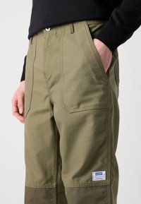 Pantalones cargo color caqui con bolsillos laterales, fabricados en un tejido duradero. Presentan costuras visibles y una etiqueta de la marca en la parte inferior de la pierna.