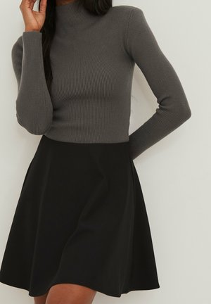 Femme portant un haut gris ajusté à manches longues côtelées et une jupe noire évasée, debout avec un bras plié et la main près du visage.