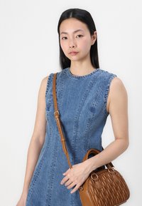 Denim jurkje zonder mouwen met een getailleerde top en stikdetails, gecombineerd met een bruine handtas van textuur met een gebogen handvat en een afneembare schouderband.