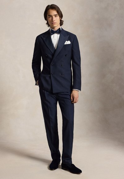 Giovane uomo in piedi che indossa un abito doppiopetto blu scuro, camicia bianca, papillon blu navy, fazzoletto bianco nel taschino e scarpe nere su sfondo semplice.