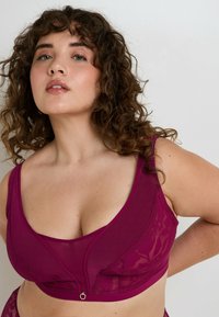 Donna dai capelli ricci che indossa un reggiseno in pizzo e rete di un magenta intenso, posando con sicurezza su uno sfondo chiaro e semplice.