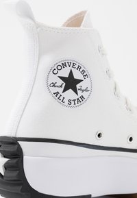 Białe trampki Chuck Taylor w wersji wysokiej z płótna, z czarną naszywką w kształcie gwiazdy, srebrnymi oczkami i teksturowaną czarną gumową podeszwą.