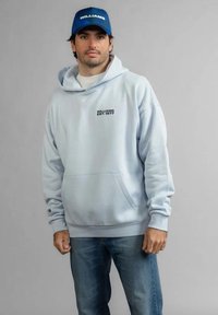 Mężczyzna w jasnoniebieskiej bluzie z kapturem z napisem "WILLIAMS EST. 1977" oraz niebieskiej czapce Williams, stojący na tle jednolitego szarego tła.