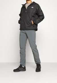 Persoon in een zwarte North Face jas, grijze broek en zwarte Adidas sneakers, staande met de handen in de zakken.