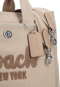 Beige canvas handbag featuring embroidered "Coach New York" text, silver hardware, and a leather tag with a chain accent.